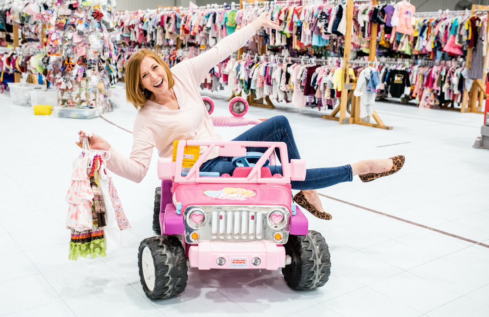 A mom sitting on toy jeep, smiling and holding children's clothing. 