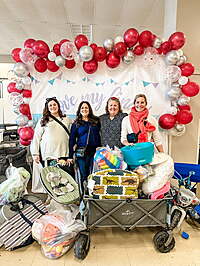 Four Moms standing in front of the JBF Red and white baloon banner. They have a wagon and lots of shopping bags that they purchased at the sale. 